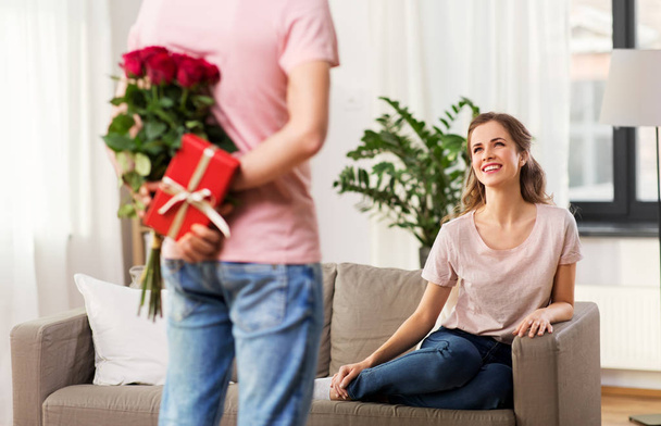 woman looking at man with flowers and gift at home - Foto, Imagem