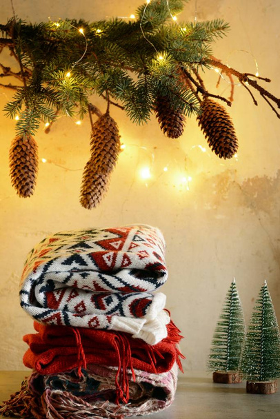 Una pila de ropa de abrigo sobre la mesa. Ambiente navideño en la casa. Habitación decorada con ramas de abeto con conos. Estilo de vida. Bodegón de otoño o invierno. Ambiente hogareño, ropa de invierno
. - Foto, Imagen
