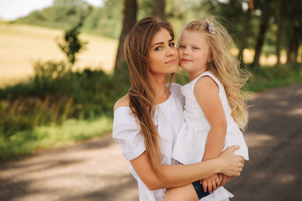 Attractive Mom and blonde hair daughter sits on road near big alley. They smile and look to natune. Front view - Foto, Imagen