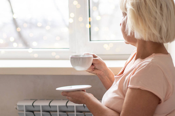 Primo piano di una donna mani che tengono una tazza di caffè calda in inverno vicino a una finestra a casa
 - Foto, immagini