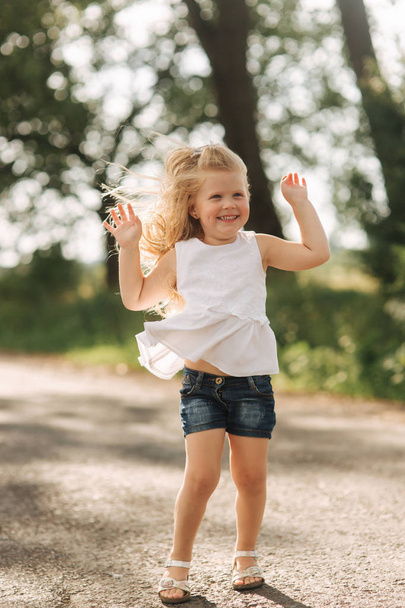 Una niña alegre y adorable con el pelo rubio baila y salta por el callejón en un cálido día de verano
 - Foto, imagen