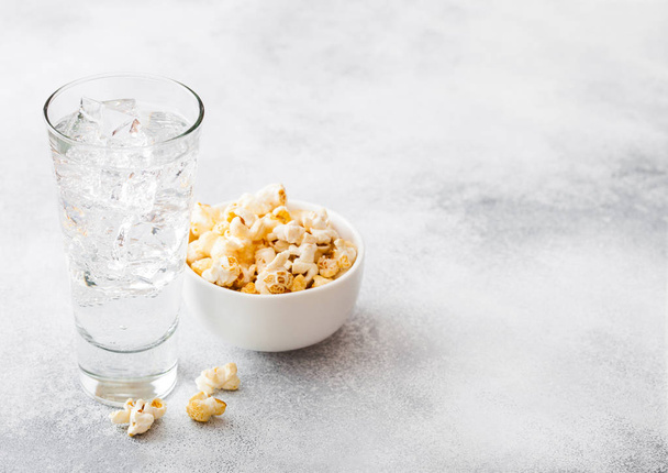 Glass of lemonade soda drink with ice cubes and whitel bowl of popcorn snack on stone kitchen background. - Fotó, kép