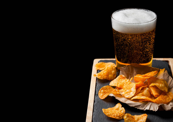 Glass of lager beer with potato crisps snack on stone board on black background. - Zdjęcie, obraz