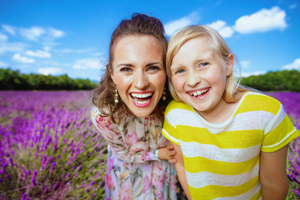 Portrait of smiling young mother and daughter against lavender field of Provence, France. Happy family portrait in blooming lavender meadow. daughter in bright yellow t-shirt. Blue midday summer sky - Photo, Image