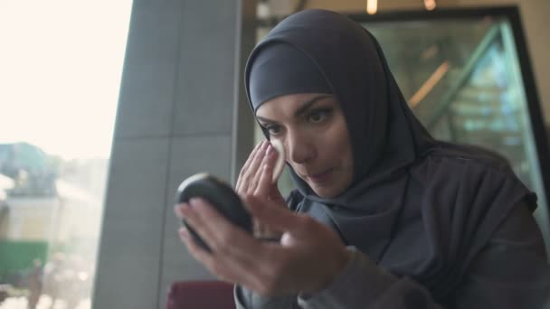 Pretty female in hijab applying powder in cafe, smiling in mirror, coquette - Imágenes, Vídeo