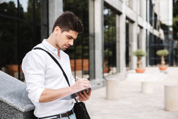 Confident successful young businessman dressed white shirt working outdoors, holding tablet, noting - Фото, изображение