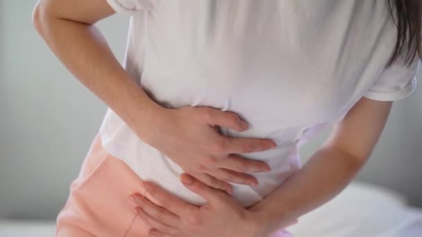 Young woman suffering from abdominal pain while sitting on bed at home - Imágenes, Vídeo