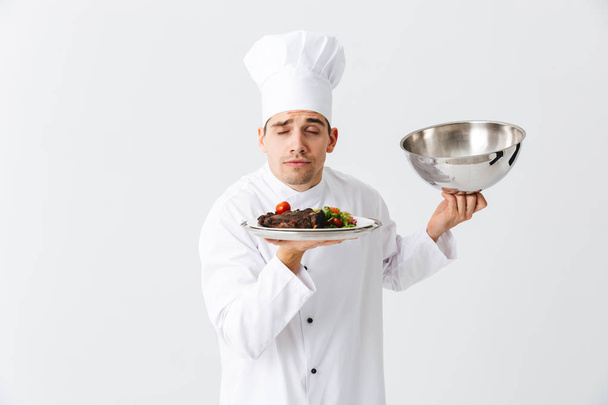 Podekscytowany mężczyzna kucharz noszenia jednolitego otwieranie pokrywy cloche na białym tle nad białym tle, Wyświetlono danie mięsne - Zdjęcie, obraz
