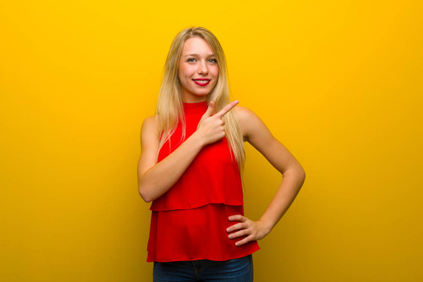 Menina com vestido vermelho sobre a parede amarela apontando para o lado para apresentar um produto
 - Foto, Imagem