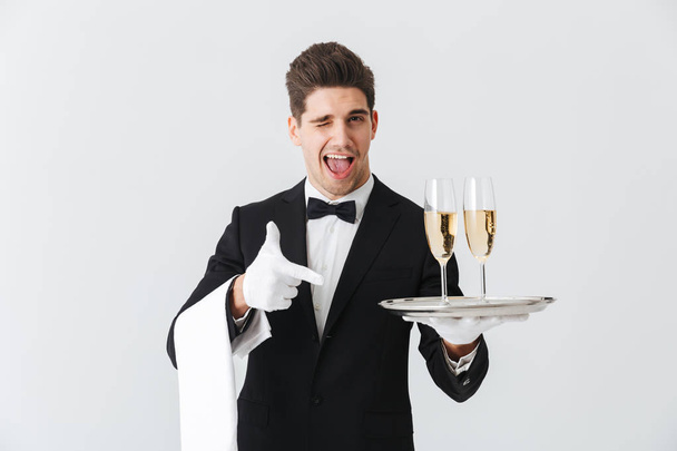 Portrait of a smiling young waiter in tuxedo offers you glass of champagne over white background - Fotoğraf, Görsel