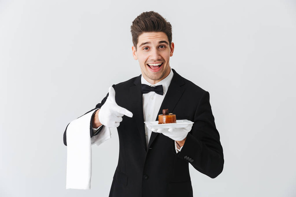 Handsome young waiter in tuxedo with bowtie holding plate with cake isolated over white background - Photo, image