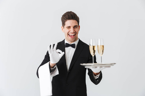 Portrait of a smiling young waiter in tuxedo offers you glass of champagne over white background - Photo, image