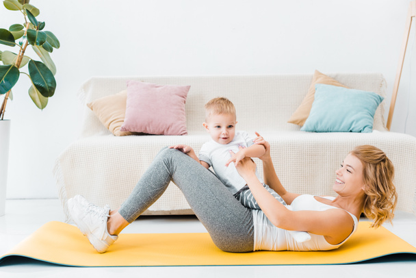 mujer sosteniendo niño pequeño hijo y entrenamiento en la alfombra de fitness en casa
 - Foto, Imagen