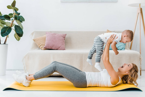 mujer acostada en la alfombra de fitness y sosteniendo lindo niño
 - Foto, Imagen