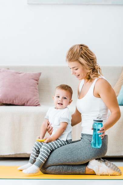 smiling cute toddler boy and attractive woman sitting on fitness mat and holding sports bottle - Foto, afbeelding
