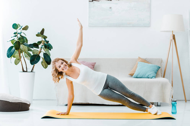 smiling woman in sportswear exercising on fitness mat and looking at camera in living room - Photo, Image