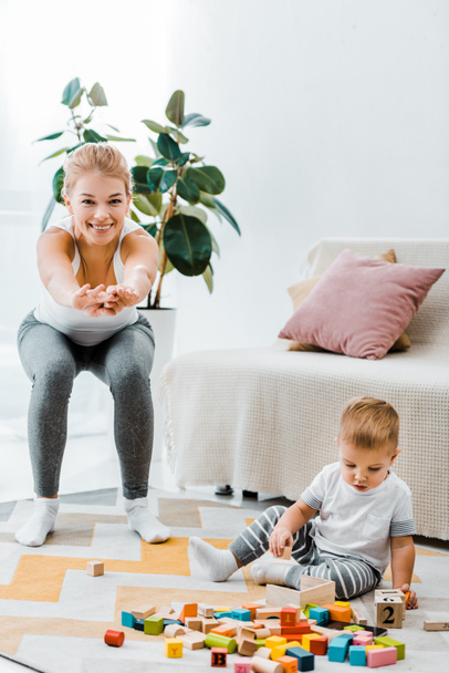 aantrekkelijke moeder doen squats en kijken camera en schattige peuter zoon zittend op een tapijt en spelen met veelkleurige kubussen in woonkamer - Foto, afbeelding