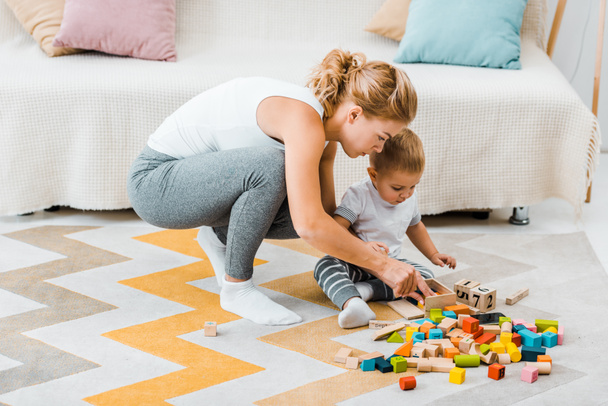 приваблива жінка і симпатичний малюк хлопчик грає з різнокольорових дерев'яних кубів на килим на дому - Фото, зображення