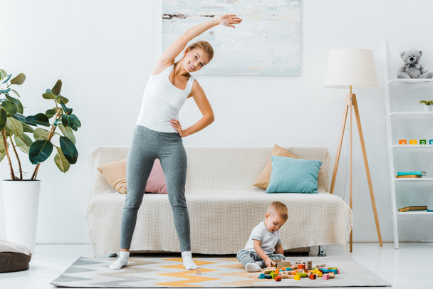 усміхнена жінка робить вправи і дивиться на камеру і малюка хлопчика, який грає з різнокольоровими кубиками на килимі у вітальні
 - Фото, зображення