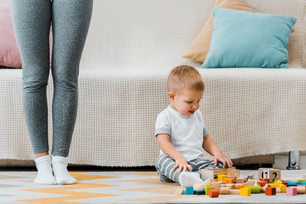 cute toddler boy playing with multicolored cubes and sitting on carpet at mom legs - Photo, Image