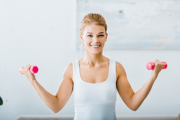 smiling woman in white sportswear doing exercise with dumbbells and looking at camera in living room - Foto, Bild