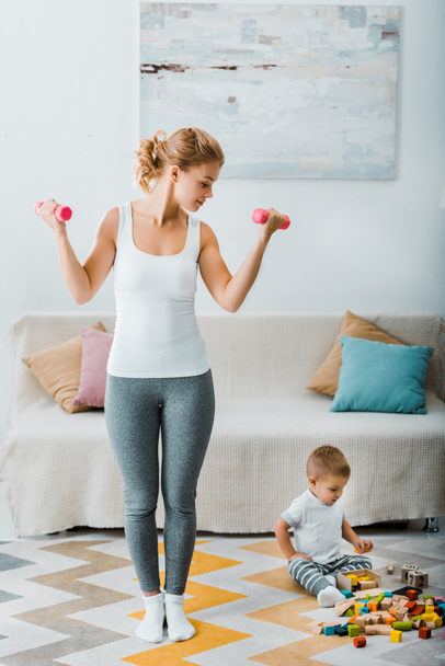 attractive woman exercising with dumbbells and looking at toddler boy sitting on carpet near multicolored cubes in living room - Photo, Image