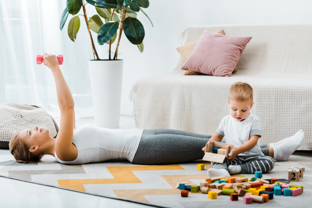 attraktive Frau, die auf Teppich liegt und mit Kurzhanteln trainiert und niedlicher Kleinkind-Junge, der Holzkiste mit bunten Würfeln im Wohnzimmer hält - Foto, Bild