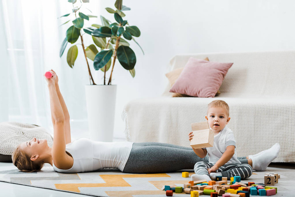 attractive woman lying on carpet and doing exercise with dumbbells and cute toddler boy holding wooden box in living room - Photo, Image
