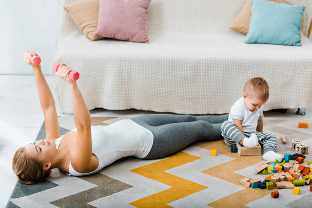 attractive woman doing exercise with dumbbells and cute toddler boy playing with multicolored wooden cubes on carpet in living room - Foto, Imagen