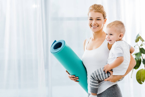 beautiful woman holding blue fitness mat and smiling cute toddler boy - Photo, Image