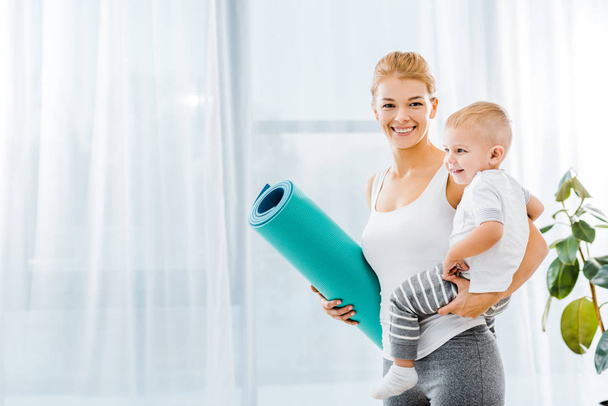 attractive woman in sportswear looking at camera, holding fitness mat and smiling toddler boy - Fotó, kép