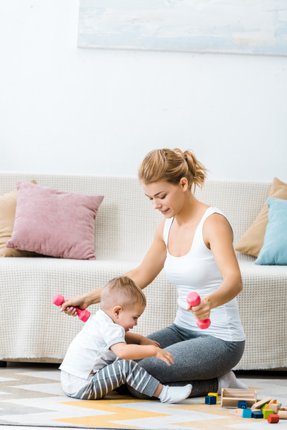 atrakcyjna kobieta trzymając hantle z toddler chłopiec siedzi na dywanie w pobliżu kanapie w salonie - Zdjęcie, obraz