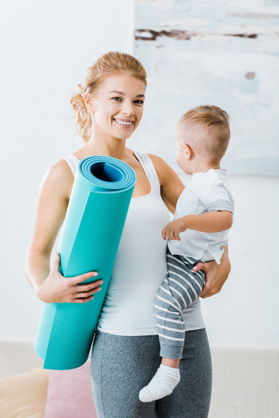 smiling woman holding fitness mat and toddler boy and looking at camera at home - Фото, изображение