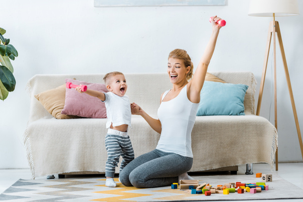 beautiful mother and cute toddler son holding dumbbells in raising hands and laughing in living room - Fotoğraf, Görsel