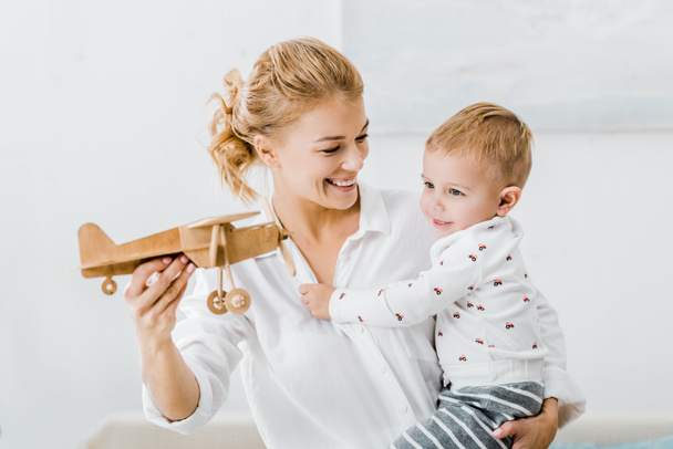 smiling woman holding wooden plane model and playing with adorable toddler boy at home - Fotó, kép