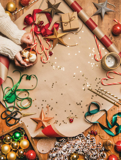 Preparing for New Year. Flat-lay of hot chocolate in mug in womans hands, candy canes, decorations, Christmas ribbons, wreath, glittering balls over gift paper background, top view, copy space - Foto, afbeelding