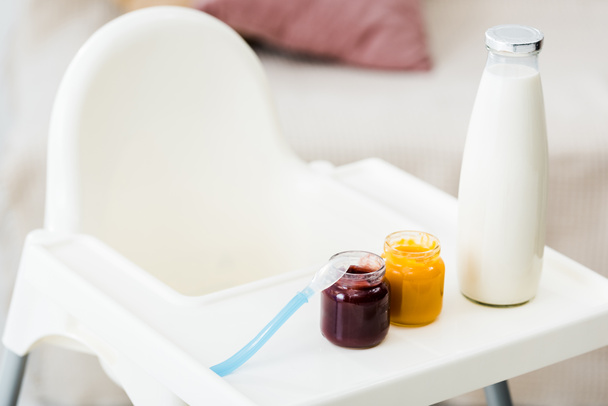 frascos de comida de bebê com colher e mamadeira de leite na cadeira alta branca na sala de estar
 - Foto, Imagem