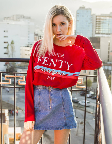 portrait of beautiful young woman in red sweater and jeans skirt posing in city - Photo, Image