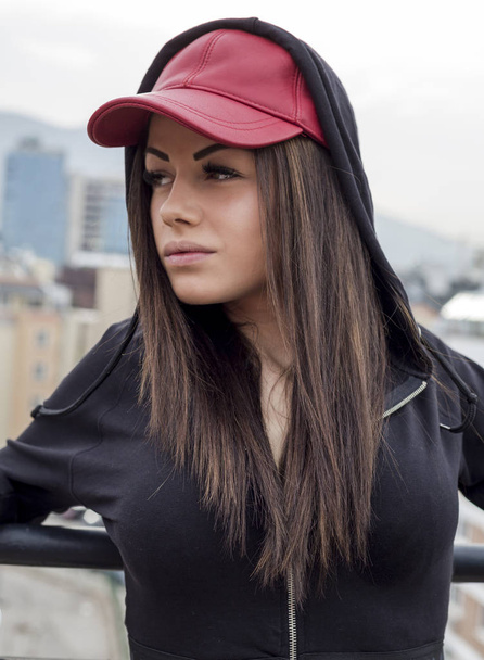 Portret van mooie jonge vrouw die zich voordeed in rode cap en zwarte hoody - Foto, afbeelding