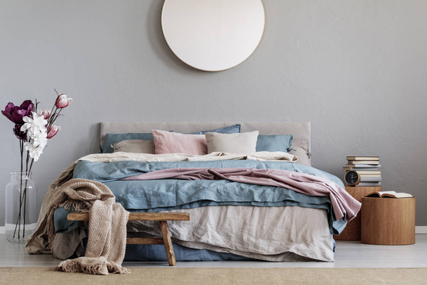 Модне кругле дзеркало на сірій стіні в шикарному інтер'єрі спальні з затишним ліжком з синім, пастельно-рожевим і бежевим листям і дерев'яними тумбочками з книгами
 - Фото, зображення