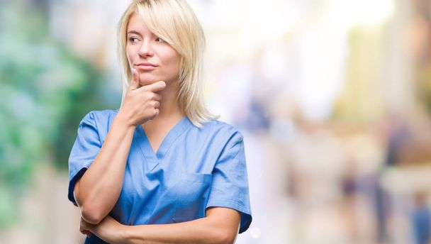 Young beautiful blonde nurse doctor woman over isolated background with hand on chin thinking about question, pensive expression. Smiling with thoughtful face. Doubt concept. - Fotoğraf, Görsel