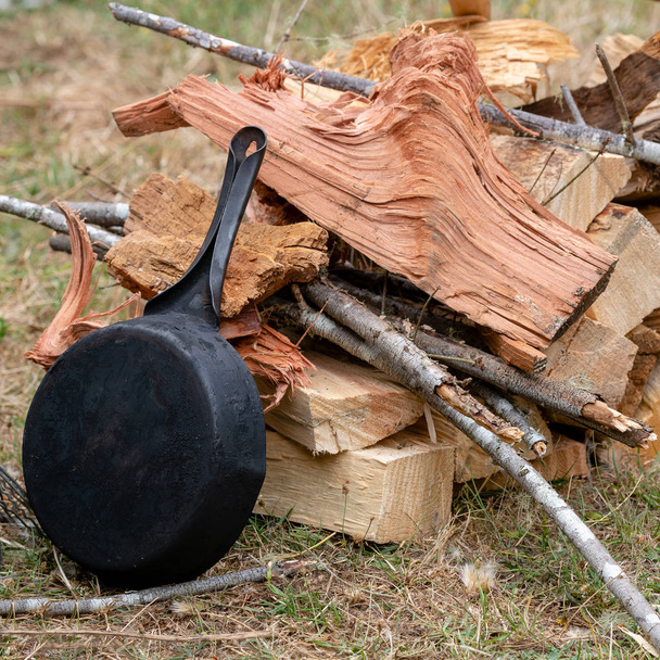 A cast iron pan outside against pile of wood suggesting a camping situation or outdoor cooking - 写真・画像