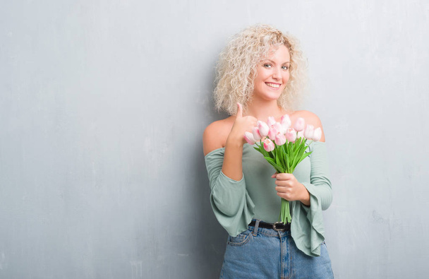 Joven mujer rubia sobre fondo gris grunge sosteniendo flores felices con una gran sonrisa haciendo signo ok, pulgar hacia arriba con los dedos, señal excelente
 - Foto, Imagen