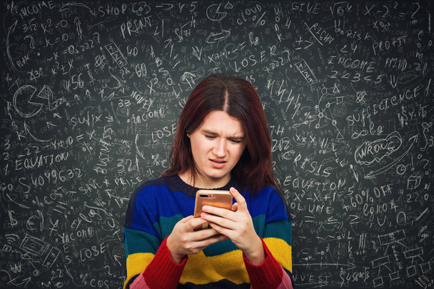 Συγχέεται νεαρή γυναίκα μπροστά από ένα μαυροπίνακα κοιτάζοντας το smartphone με αμηχανία πρόσωπο συναίσθημα που προσπαθεί να λύσει το δύσκολο μαθηματικά υπολογισμός, τύπος και εξισώσεις. Νέες τεχνολογίες στην εκπαίδευση. - Φωτογραφία, εικόνα