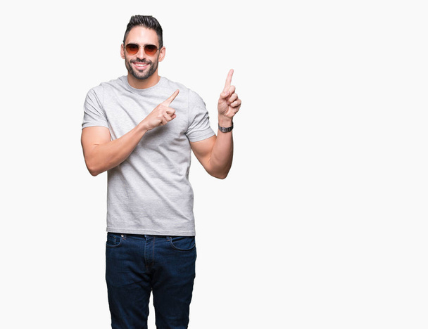 Jonge knappe man dragen van een zonnebril op geïsoleerde achtergrond glimlachen en kijken naar de camera met twee handen en vingers wijzen naar de kant. - Foto, afbeelding