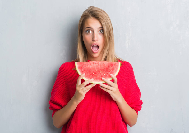 Beautiful young woman over grunge grey wall eating water melon scared in shock with a surprise face, afraid and excited with fear expression - Фото, изображение