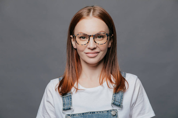 Happy young woman with glad facial expression, has healthy skin, wears optical glasses, white t shirt and denim sarafan, models over grey background, going to meet with friends, poses indoor - Photo, Image