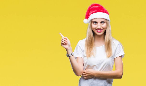 junge schöne blonde Frau Weihnachtsmütze über isoliertem Hintergrund mit einem breiten Lächeln im Gesicht, zeigt mit Hand und Finger zur Seite und schaut in die Kamera. - Foto, Bild