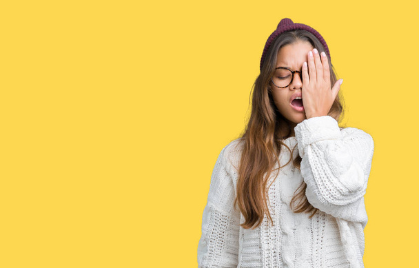 Young beautiful brunette hipster woman wearing glasses and winter hat over isolated background Yawning tired covering half face, eye and mouth with hand. Face hurts in pain. - Photo, Image