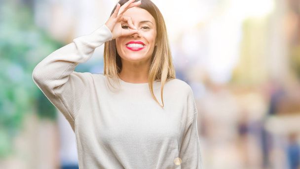 Молодая красивая женщина случайный белый свитер на изолированном фоне делает хорошо жест с улыбкой на руке, глаза смотрят сквозь пальцы с счастливым лицом
. - Фото, изображение